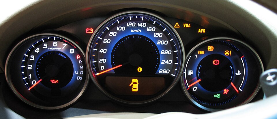 2006 photo of a softly glowing car dashboard. The prominent gauges are a tachometer on the left that includes a gear indicator, a speedometer displaying km/h in the center, and fuel and coolant gauges on the right. Most of the indicator lights are on.