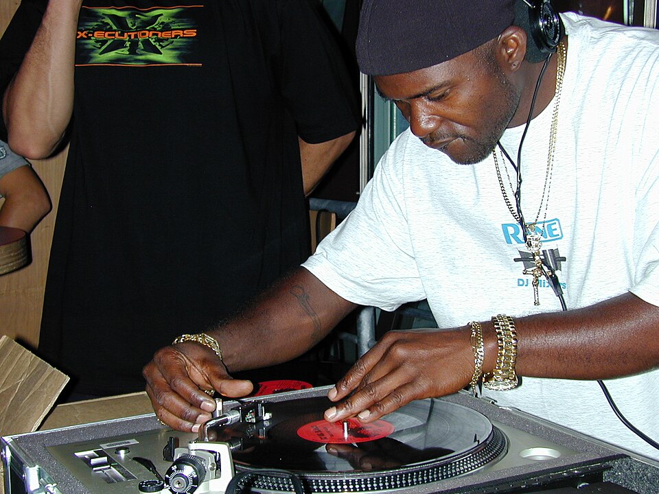 2002 photo of NYC-based DJ Grand Wizzard Theodore demonstrating the now ubiquitous DJ scratch technique that he invented in 1975. Theodore has brown skin and is wearing a black durag, light gray t-shirt, headphones, and gold jewelry. He is intently working a turntable.