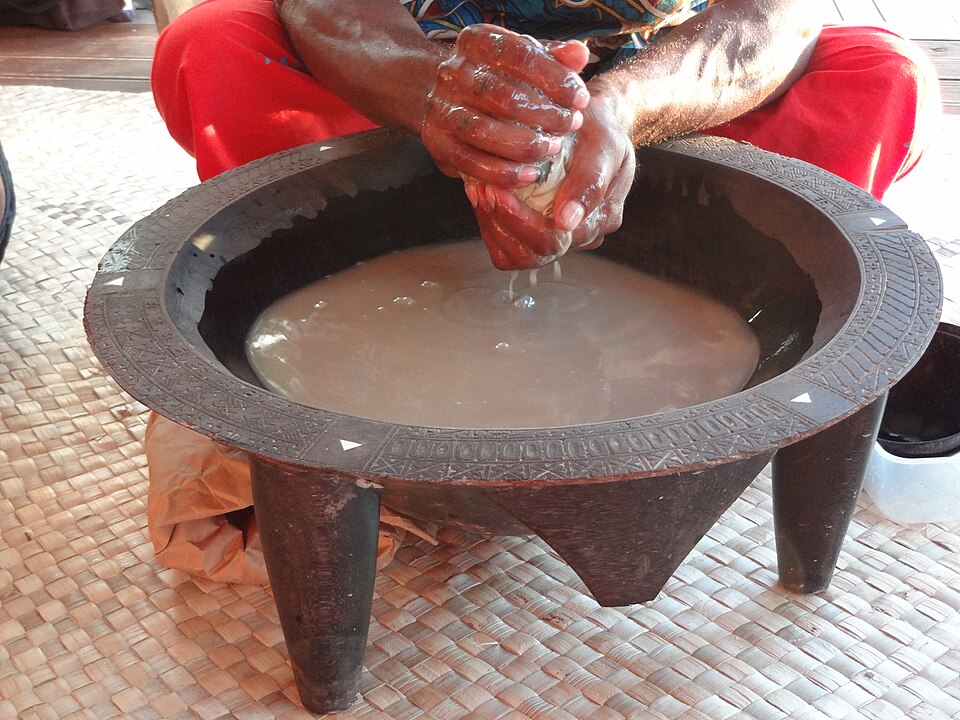 Photo of a traditional Fijian method of kava preparation. The focus is a large metal urn of brown liquid on an outdoor rattan floor. A sitting person with brown skin and red pants, face out of frame, mixes a drink vessel above the urn.