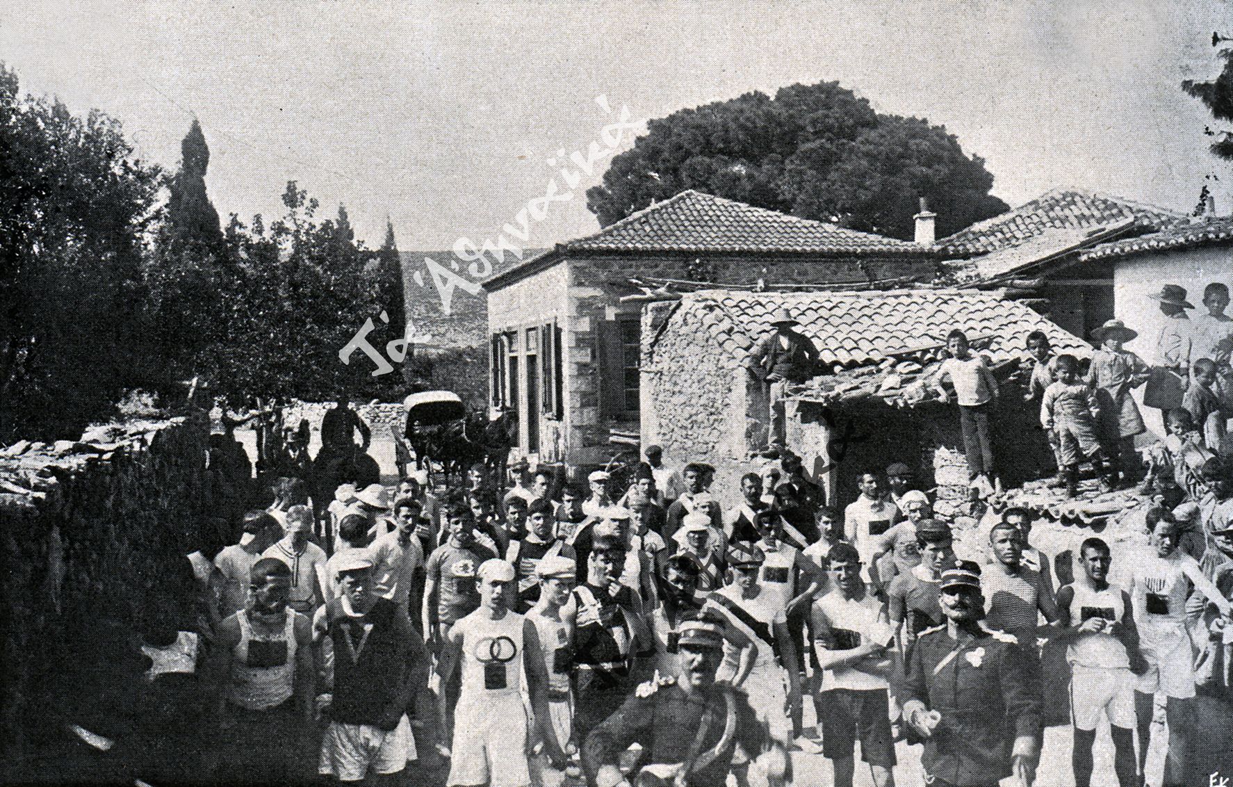 Photo of the start of the 1896 Olympic marathon, in the Greek town of Marathon. About 50 men and several horses are lined up on a road in front of a large one-story building in the countryside. 8 children watch from the roof of the building.