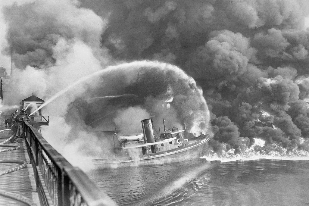 Black-and-white photo of the Cuyahoga River on fire. Most of the frame is filled with smoke. Firefighters on a bridge on the left spray water at the blaze from hoses.