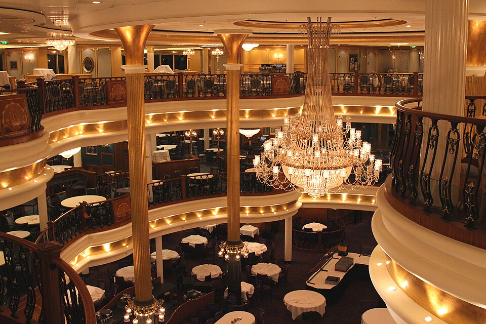 Photo of a fancy-schmancy multi-story dining room aboard a cruise ship. A central chandelier warmly lights the many elegant balconies and tablecloths.