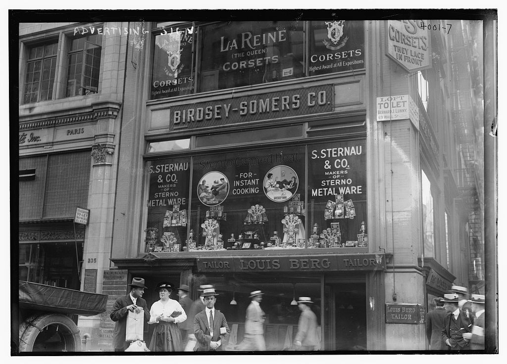 Late 1910s photo of the NYC storefront at 27th St and 5th Ave. The second floor of the busy storefront is labeled Birdsey-Somers Co. and filled with an advertising display for Sterno Canned Heat and metalware, sold by S. Sternau & Co. The first floor is occupied by Louis Berg, Tailor, while La Reine (The Queen) Corsets takes up the third.