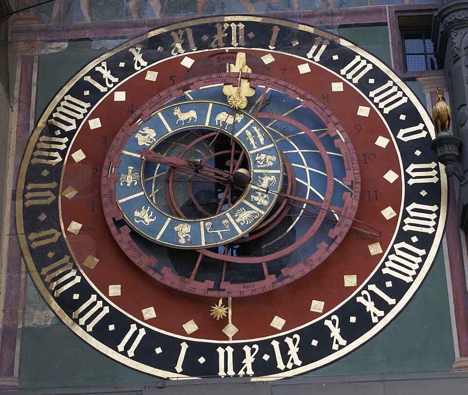 Photograph of an astronomical clock in Switzerland built perhaps around 1405. The clock only has an hour hand and Roman numerals from 1 to 12 twice going clockwise. Within the clock face is a smaller dial with the 12 zodiac animals and clockwork machinery in stark red, blue, and gold.